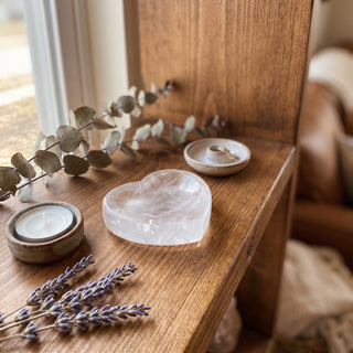 Clear Quartz Heart Bowl displayed on a wooden home altar shelf beside a tealight candle, shown at true size in warm natural light.