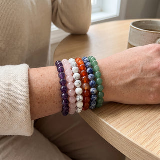 The six intention bracelets from the display kit worn stacked on a wrist in natural light: amethyst, rose quartz, howlite, carnelian, sodalite and green aventurine 8mm beads.