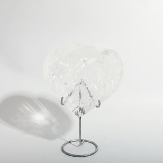 Clear Quartz Heart on Stand Unique #2, a polished clear quartz heart on a silver metal stand showing internal veils and rainbow inclusions, photographed on a clean white background. Stonebridge Imports.