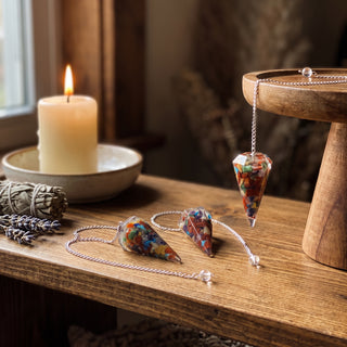 Chakra Orgone Pendulum 6 Facet & Bead - 3 Pack on a wooden altar shelf beside a candle, three multicoloured chakra orgone pendulums at true size in warm natural light.
