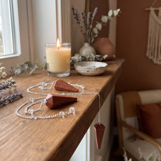 Brown Goldstone Pendulum 6 Facet & Bead - 3 Pack on a wooden altar shelf beside a candle, three red-brown sparkling goldstone pendulums at true size in warm natural light.