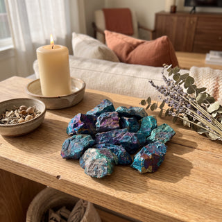 Rough chalcopyrite peacock ore chips poured in a loose heap on a wooden home altar shelf beside a candle, shown at true size in warm natural light.