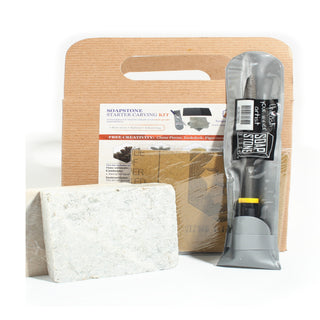 Soapstone Starter Carving Kit showing its two pale grey-green soapstone blocks, packaged steel rasp tool and kraft carry pack, photographed on a clean white background. Stonebridge Imports.