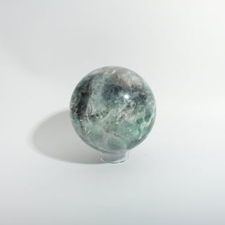 Fluorite Green and Purple Sphere Unique #11, showing green and grey fluorite with white feathering and dark inclusions, photographed on a clean white background. Stonebridge Imports.