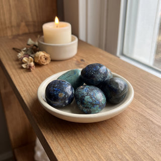 Azurite Tumbled Stones heaped in a small ceramic dish on a wooden home altar shelf beside a candle, shown at true size in warm natural light.