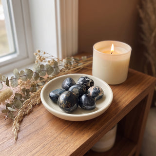 Covellite Tumbled Stones heaped in a small ceramic dish on a wooden home altar shelf beside a candle, shown at true size in warm natural light.