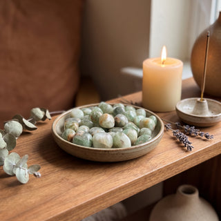 Green Fluorite Tumbled Stones - India heaped in a small ceramic dish on a wooden home altar shelf beside a candle, shown at true size in warm natural light.
