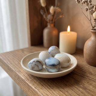 Blue Opal Tumbled Stones heaped in a small ceramic dish on a wooden home altar shelf beside a candle, shown at true size in warm natural light.