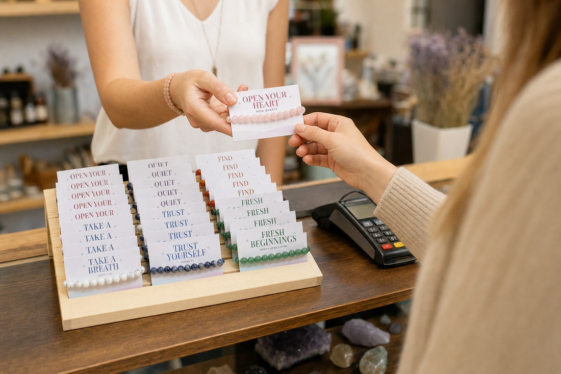 A shop owner hands a carded rose quartz bracelet to a customer, the Intention Bracelet Display Kit on the counter by the card terminal.