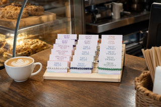 The Intention Bracelet Display Kit beside the till of a cozy coffee shop next to a latte, 30 carded crystal bracelets ready to sell.